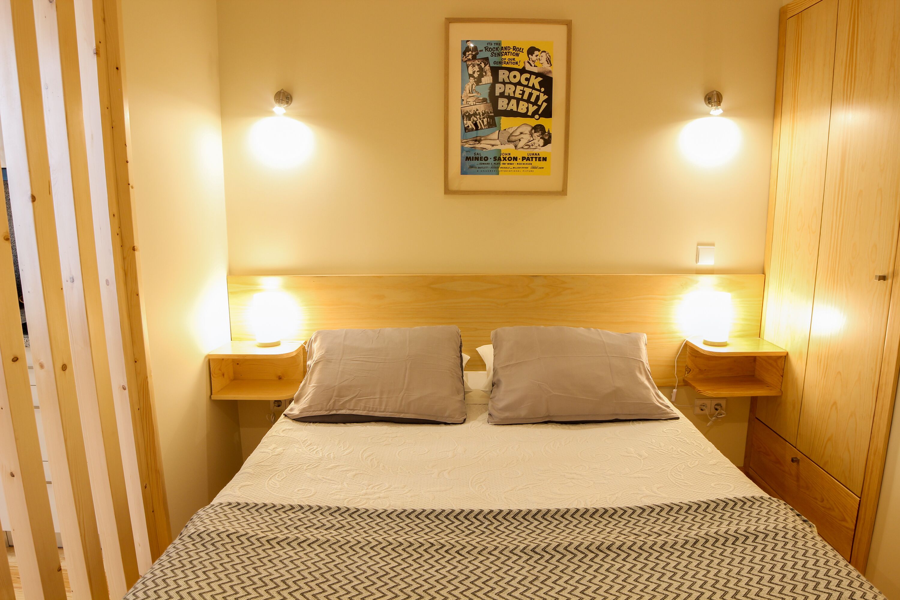 A neatly made bed with two pillows, a wooden headboard, bedside lamps, and a poster on the wall.