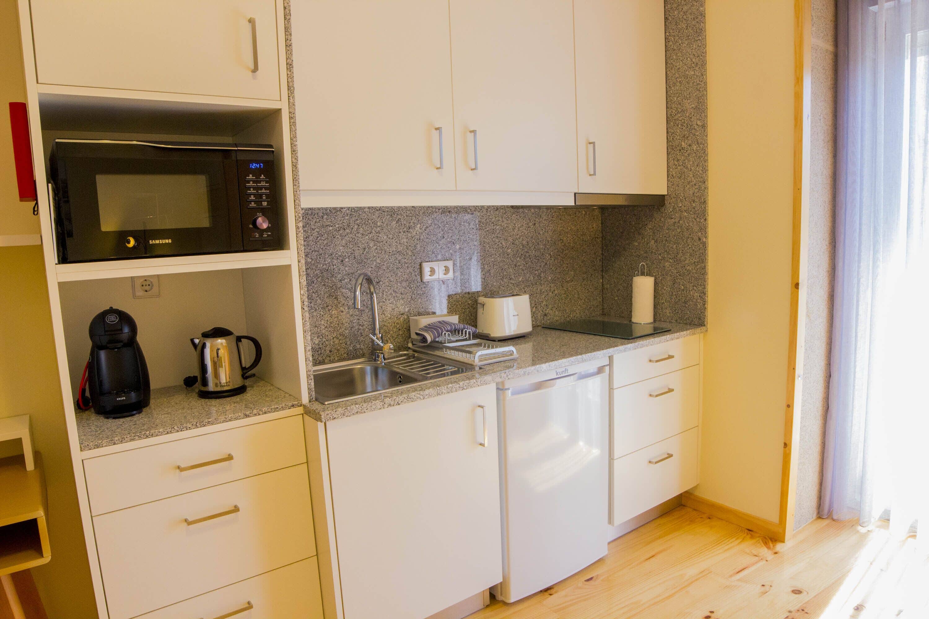A compact kitchen with white cabinets, a microwave, a coffee maker, a kettle, and a dishwasher.