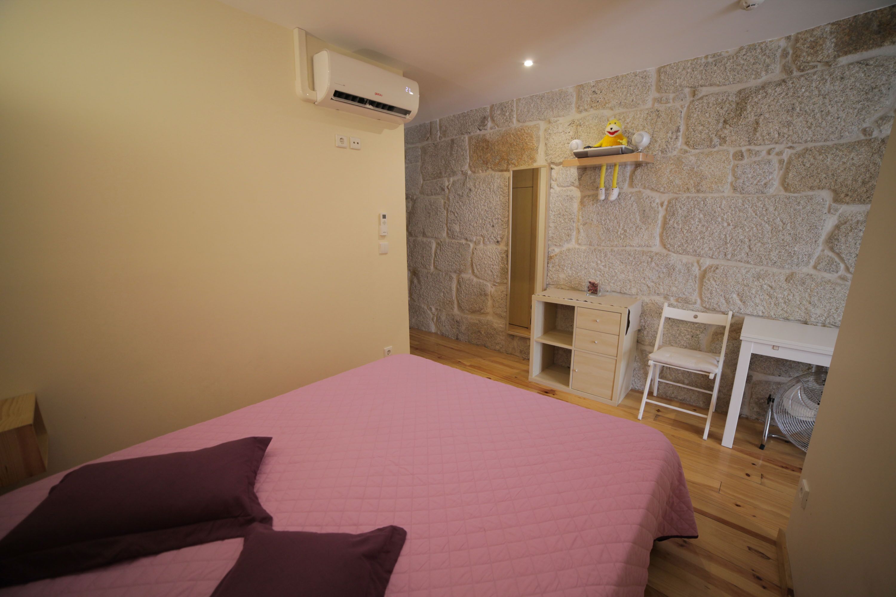 A bedroom with a pink bed, a wooden desk, a chair, and a stone wall.