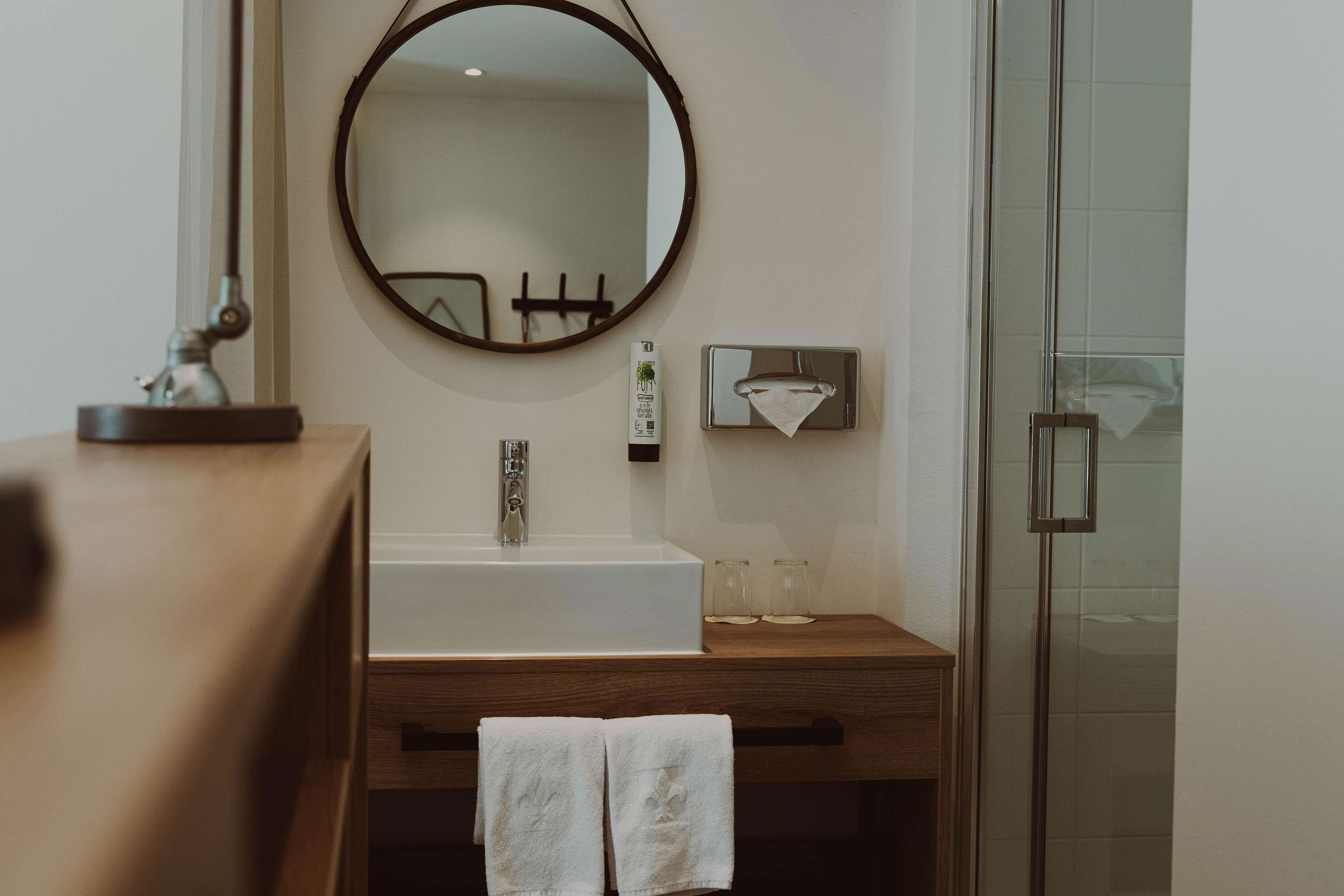 Une salle de bain avec un meuble-lavabo en bois, un miroir rond, un lavabo blanc et une cabine de douche en verre.