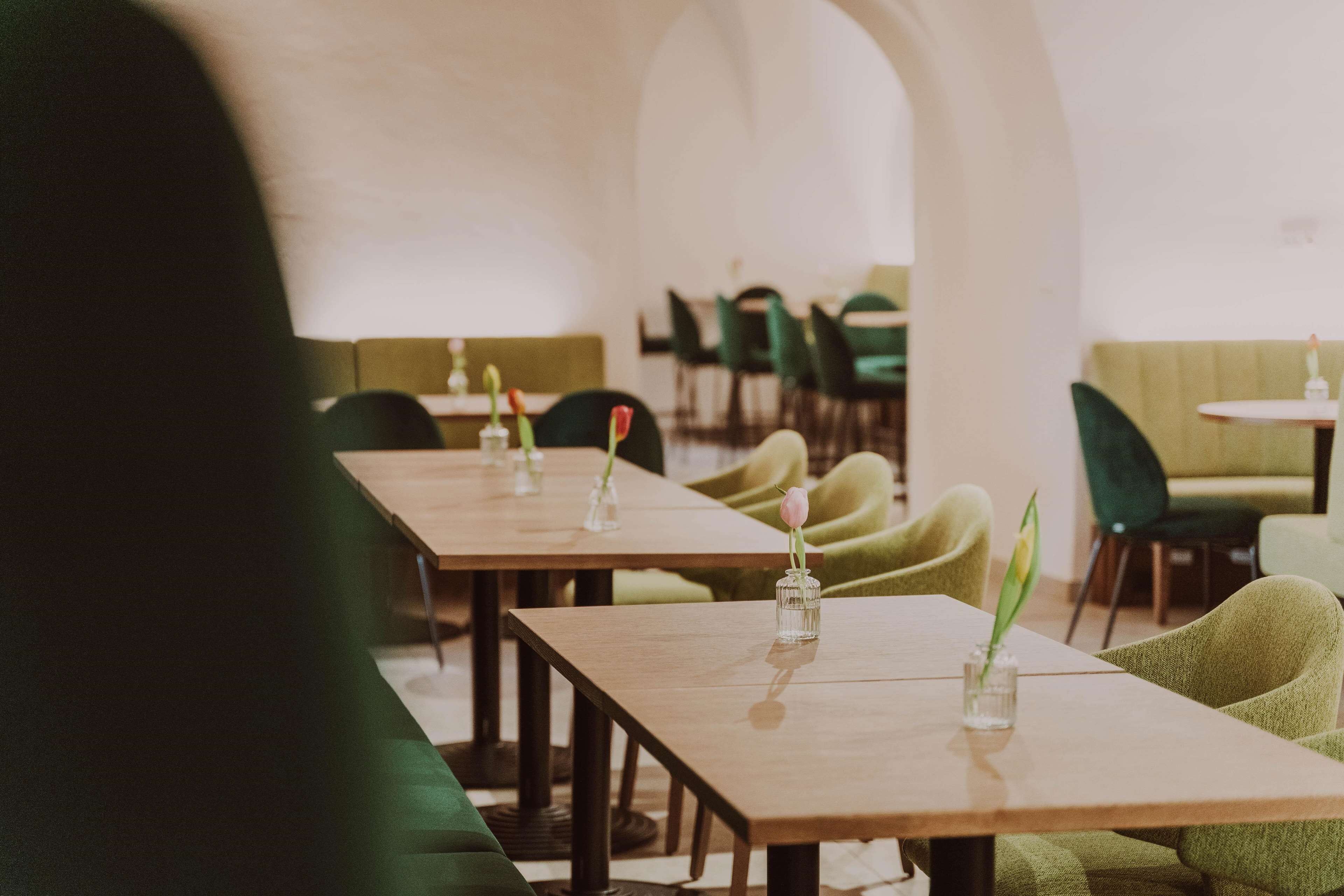Un restaurante con mesas de madera largas, sillas tapizadas de verde y jarrones con flores.