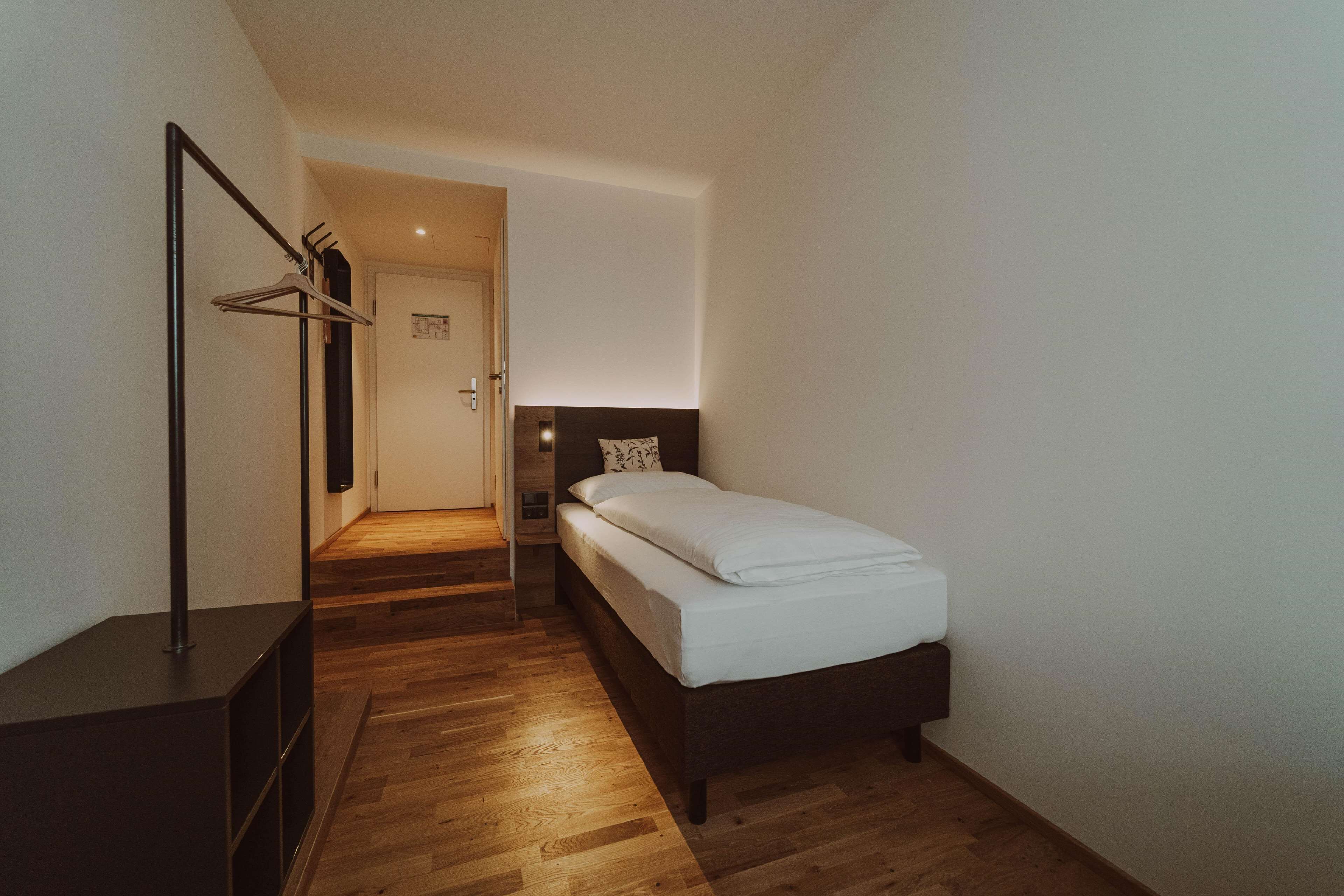 A bedroom with a bed, a wooden floor, a coat rack, and a dark-colored dresser.