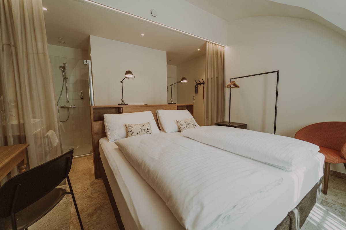 A hotel room with a large bed, two bedside tables, a desk, a chair, a shower area, and a mirror.