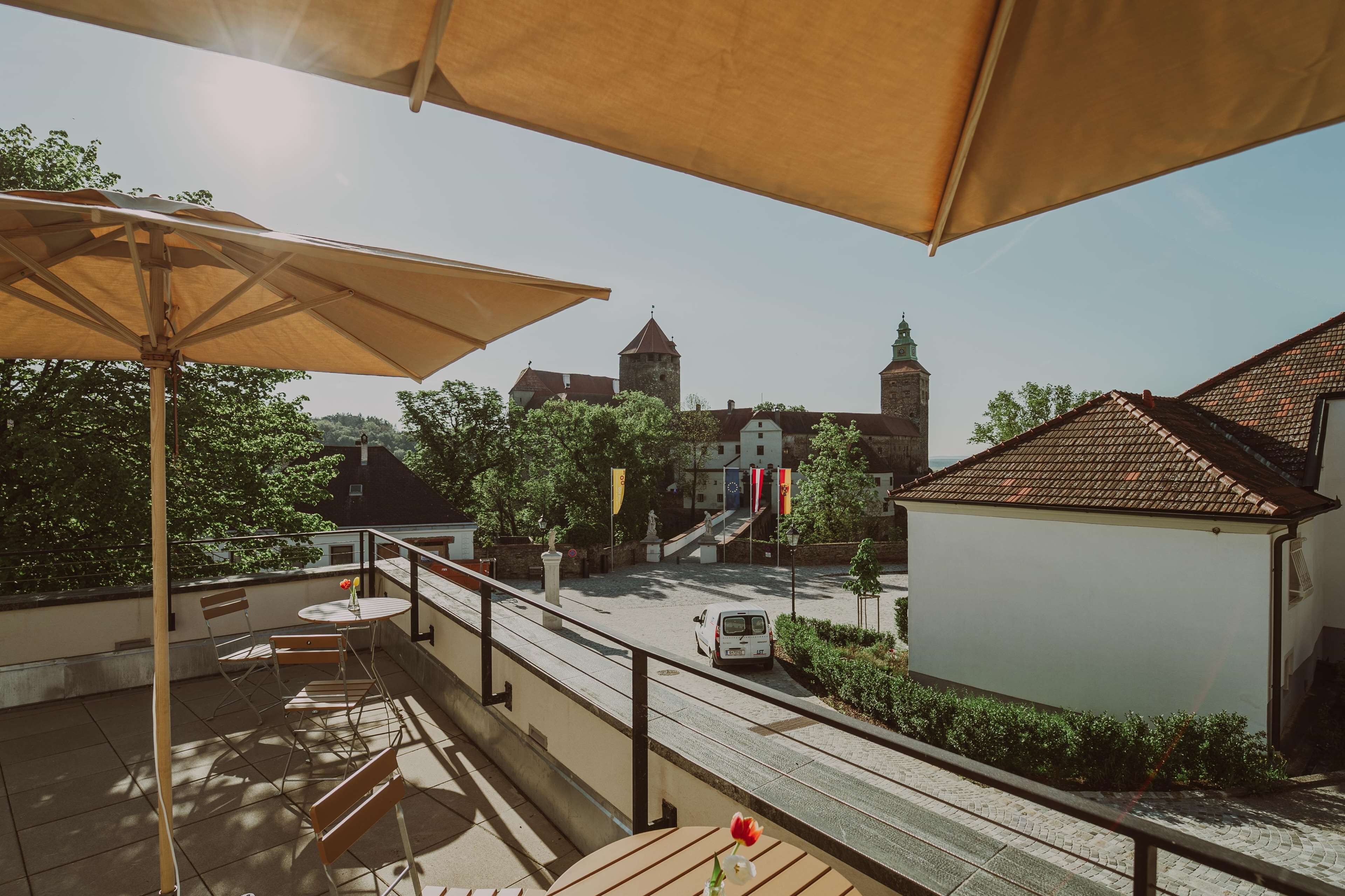 Une terrasse extérieure avec une table et des chaises, un grand parasol, et une vue sur une rue bordée d’immeubles et d’une tour en arrière-plan.