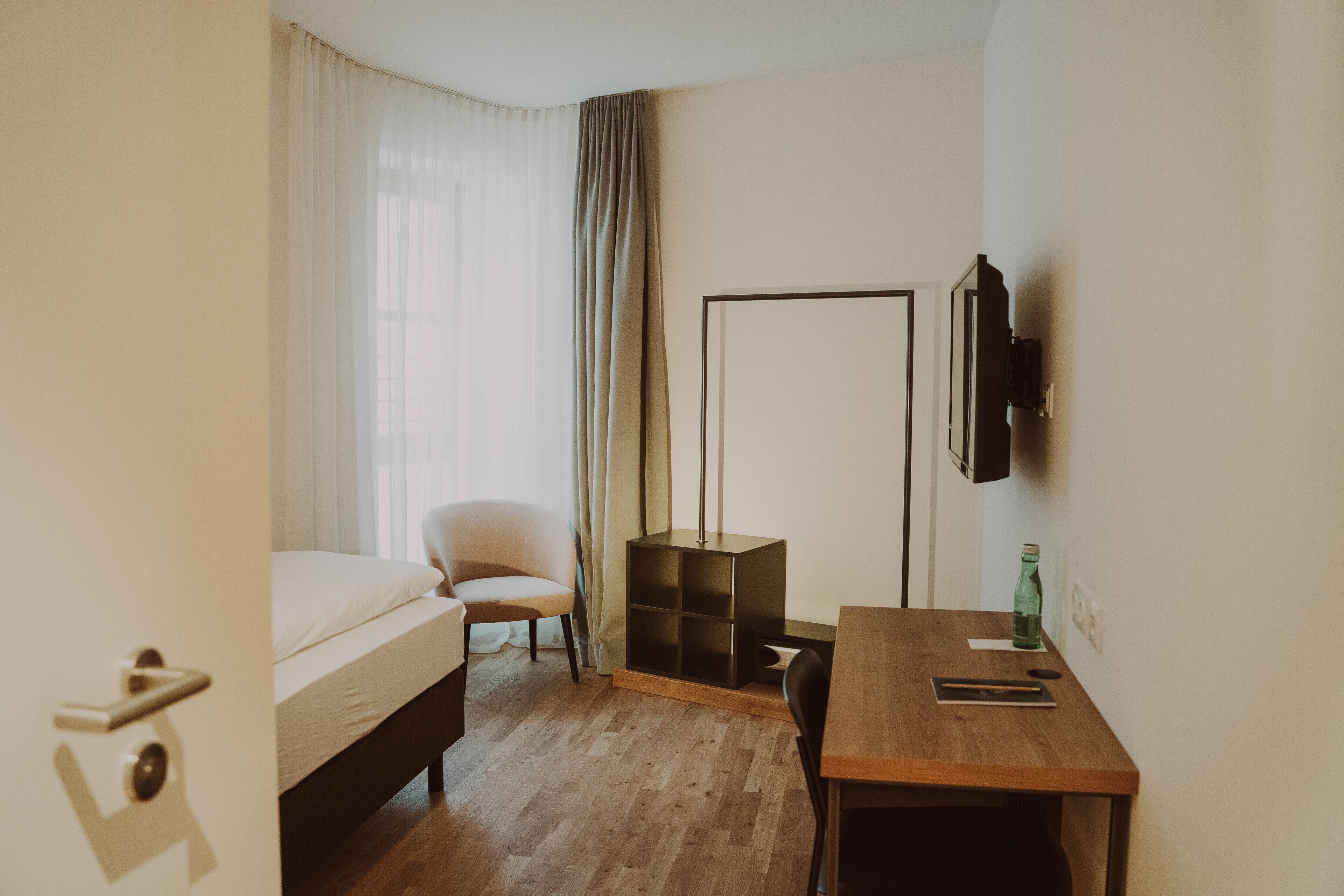 Habitación de hotel con cama, una silla, escritorio y televisor montado en la pared.