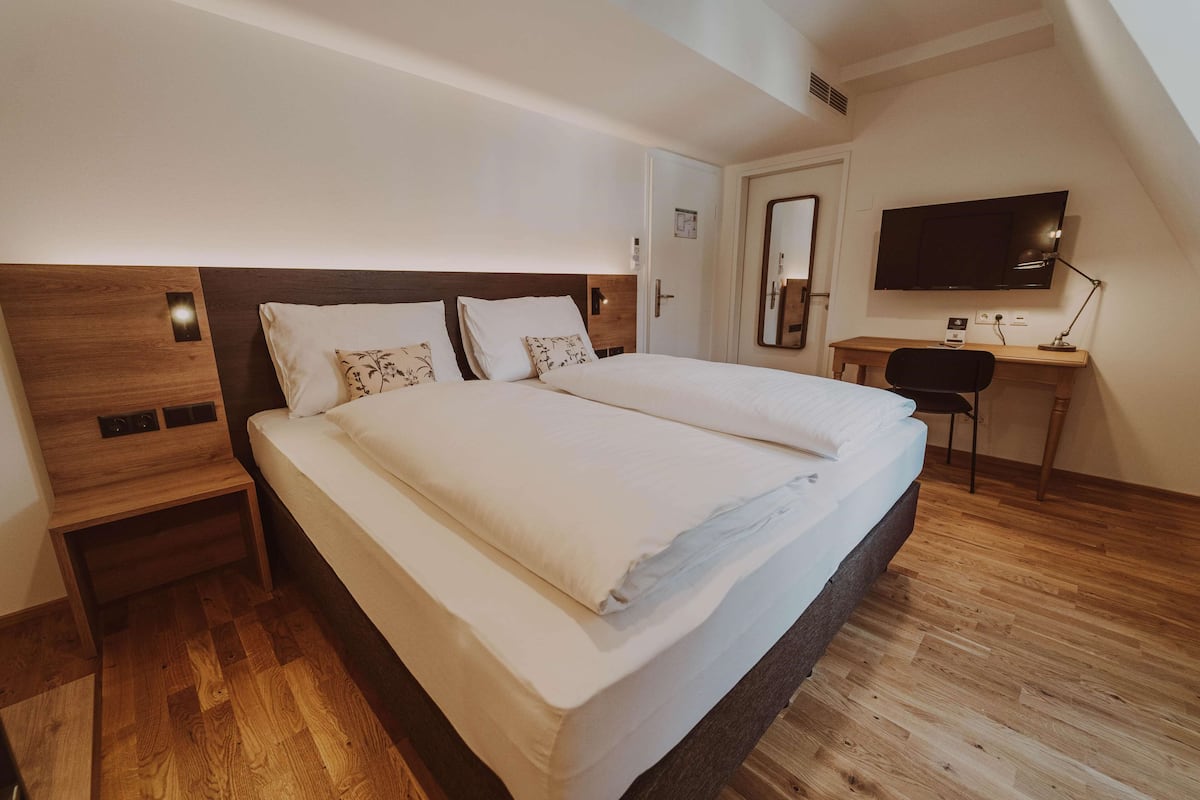 A modern hotel room with a large bed, wooden headboard, a desk with a chair, a flat-screen TV, and a mirror on the wall.