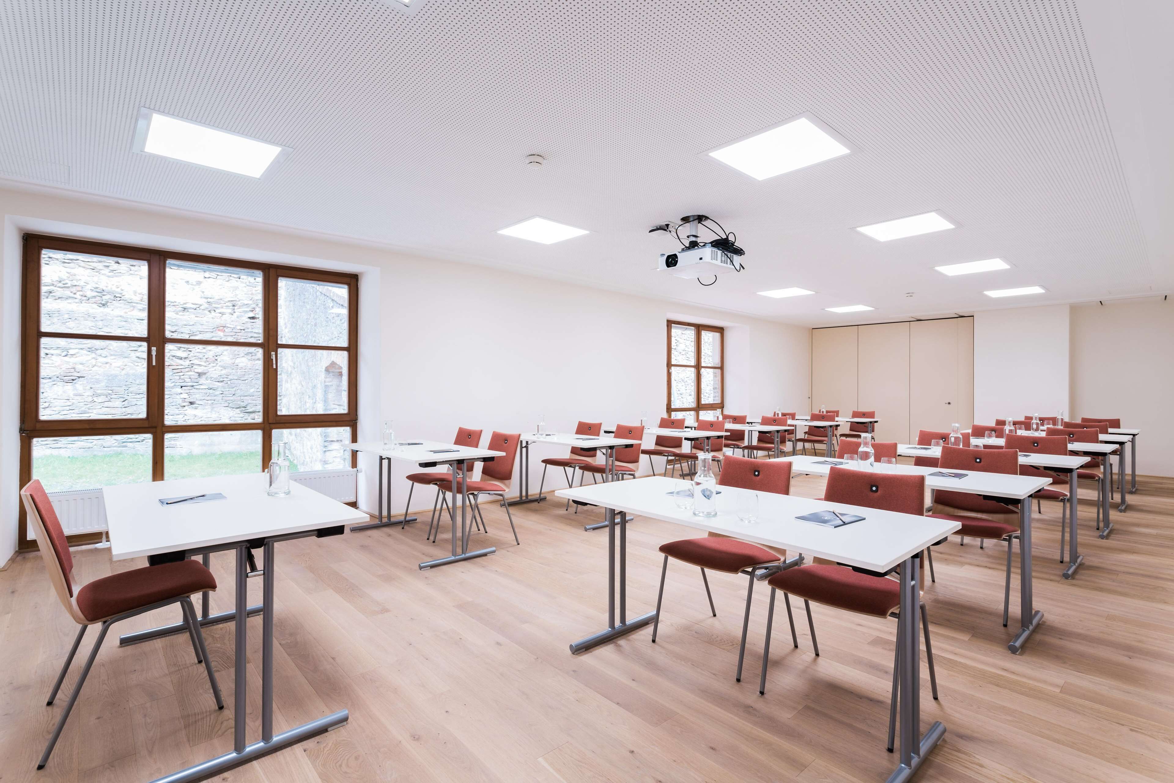 Un aula con mesas blancas y sillas rojas, ventanas amplias y proyectores de techo.