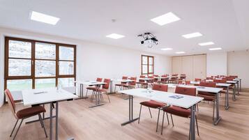 Un aula con mesas blancas y sillas rojas, ventanas amplias y proyectores de techo.