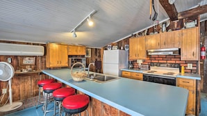 A kitchen with wooden cabinets, a central island with stools, a refrigerator, and a stove. - Waterfront Glen Lake Cabin w/ Boat Dock & Deck (Queensbury)