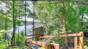 A wooden deck with a metal railing overlooking a lake, surrounded by dense trees. - Waterfront Glen Lake Cabin w/ Boat Dock & Deck (Queensbury)
