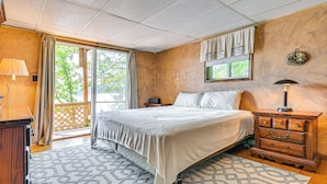 A bedroom with a bed, a dresser, a nightstand, a lamp, a window with curtains, and a view of greenery outside. - Waterfront Glen Lake Cabin w/ Boat Dock & Deck (Queensbury)