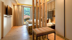 A modern hotel room with a wooden floor, a bed with a headboard, a bench, a TV mounted on the wall, and a view of a green landscape through a window. - Alpin Apartments (Ramsau im Zillertal)