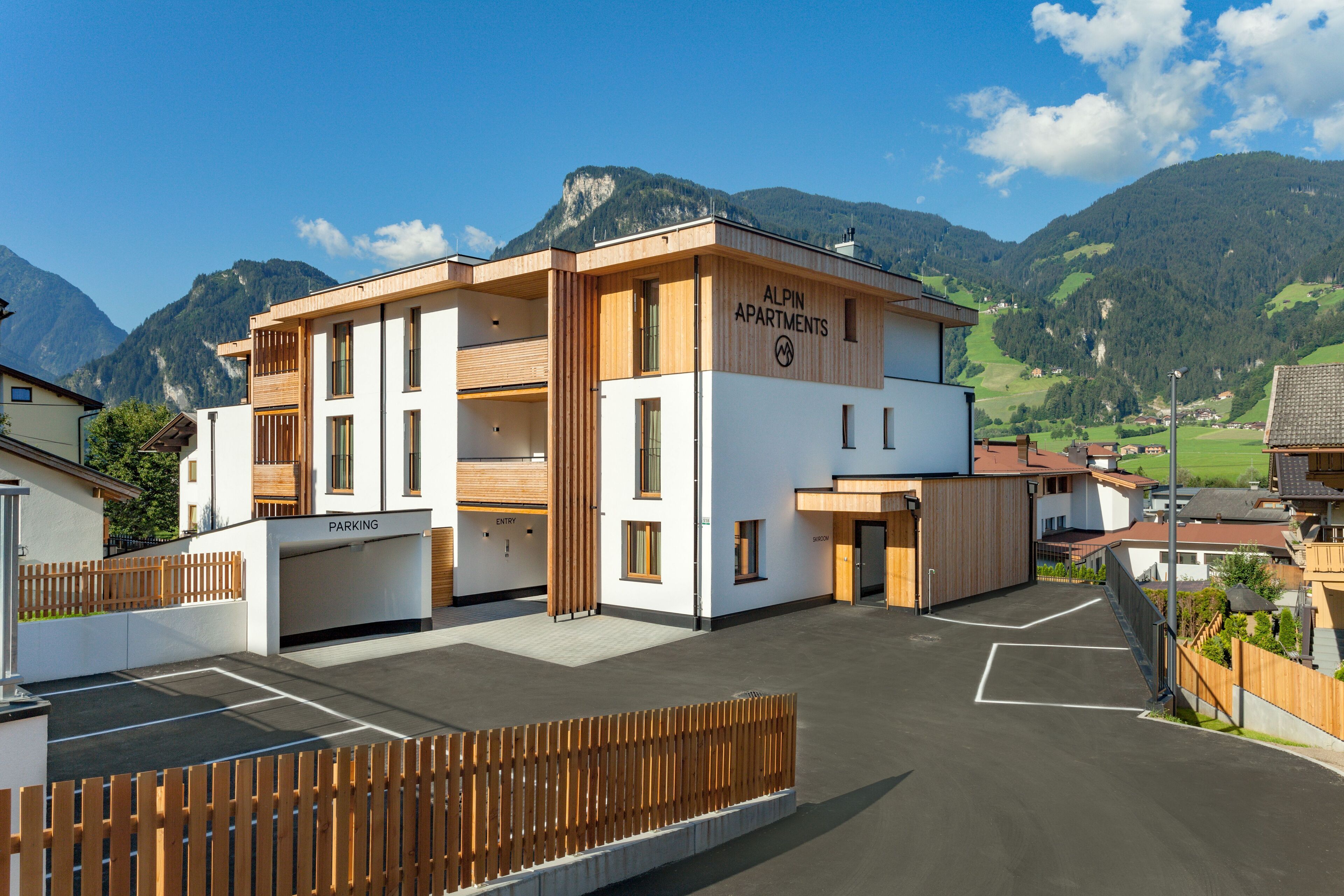 A modern building with a sign reading "ALPIN APARTMENTS" and a parking area in front.