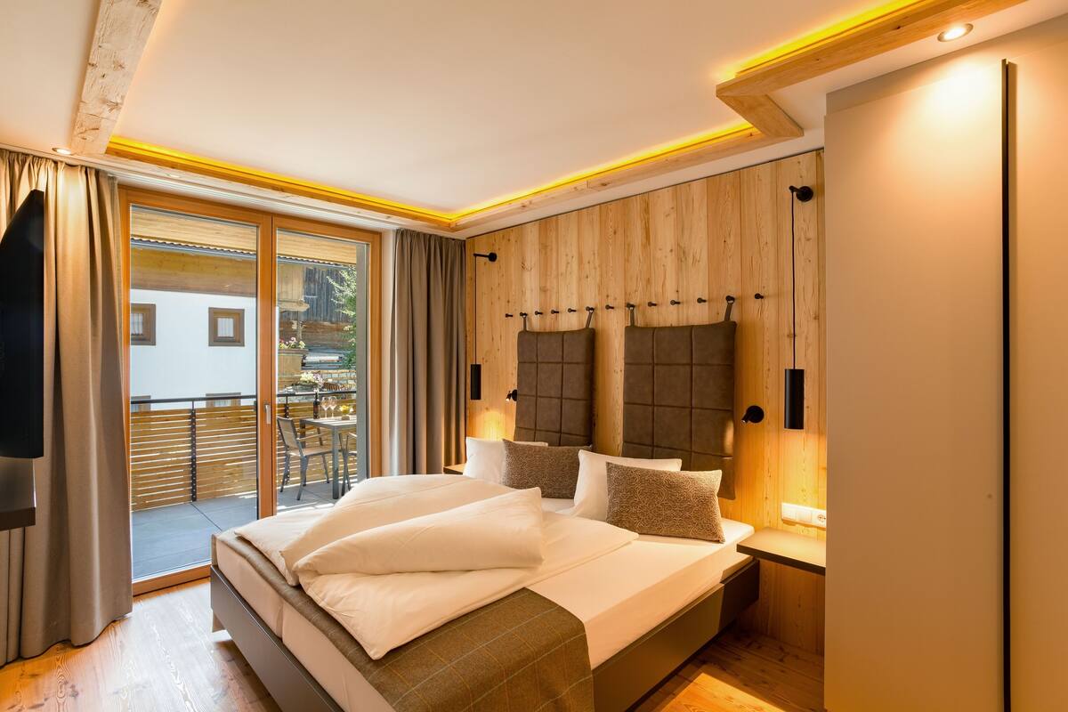 A modern bedroom with a large bed, wooden panel walls, and a balcony with outdoor seating.
