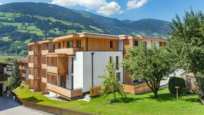 A modern multi-story building with wooden paneling, a white facade, and a view of green hills and mountains in the background. - Alpin Apartments (Ramsau im Zillertal)