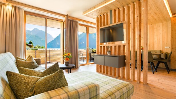 A modern hotel room with a large bed, a TV mounted on a wooden wall, a balcony with a view of mountains, and a dining area. - Alpin Apartments (Ramsau im Zillertal)