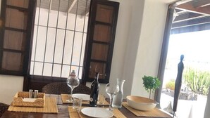 A dining table set with plates, glasses, and a bottle, with a window and plants in the background. - Villa Mare Bella Ciao Sea view private pool A / C 500m from the beach (Puerto del Carmen)