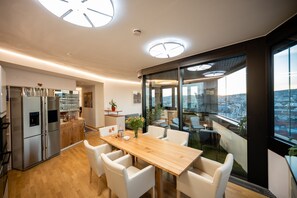 Une cuisine moderne avec une table en bois et des chaises blanches, un réfrigérateur encastré et une vue sur la ville grâce à de grandes fenêtres.
