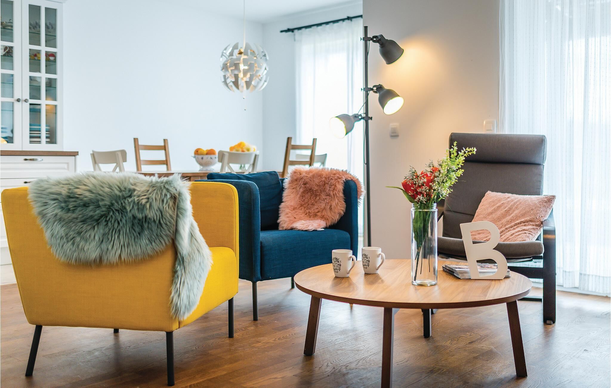 Ein modernes Wohnzimmer mit einem gelben Sessel, einem blauen Sofa und einem grauen Sessel. Ein runder Couchtisch mit zwei Tassen und einer Vase mit Blumen.
