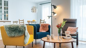 Una sala moderna con un sillón amarillo, un sofá azul y un sillón gris. Hay una mesa de centro redonda de madera con dos tazas y un florero con flores.