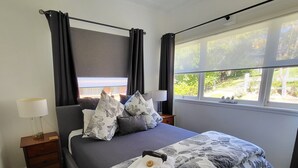 A neatly made bed with floral bedding, a bedside table with a lamp, and a window with a view of greenery. - Winston Cottage Leura - BEST Location & Views (Leura)