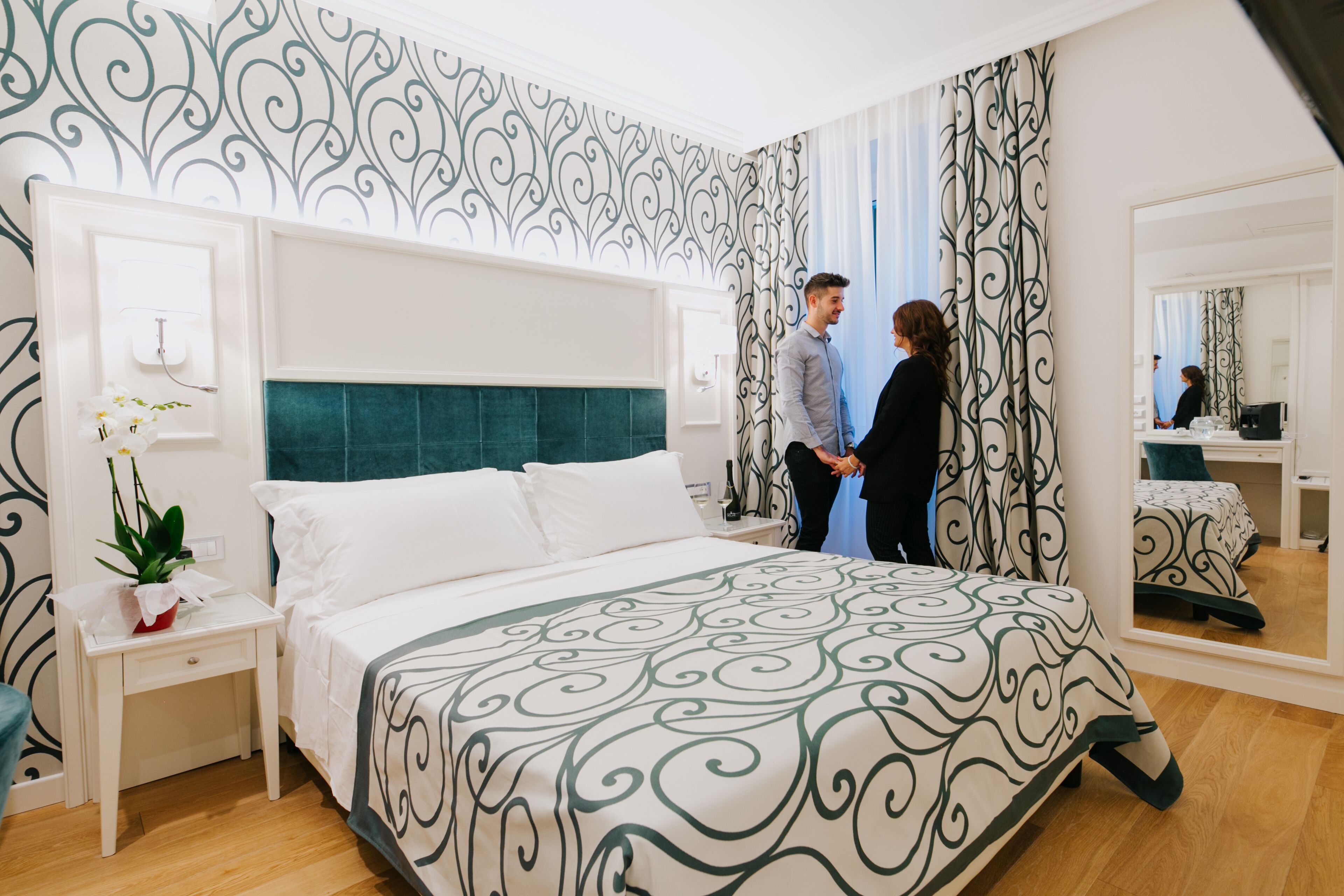 A hotel room with a large bed, a bedside table with a plant, a mirror, and a patterned wall.