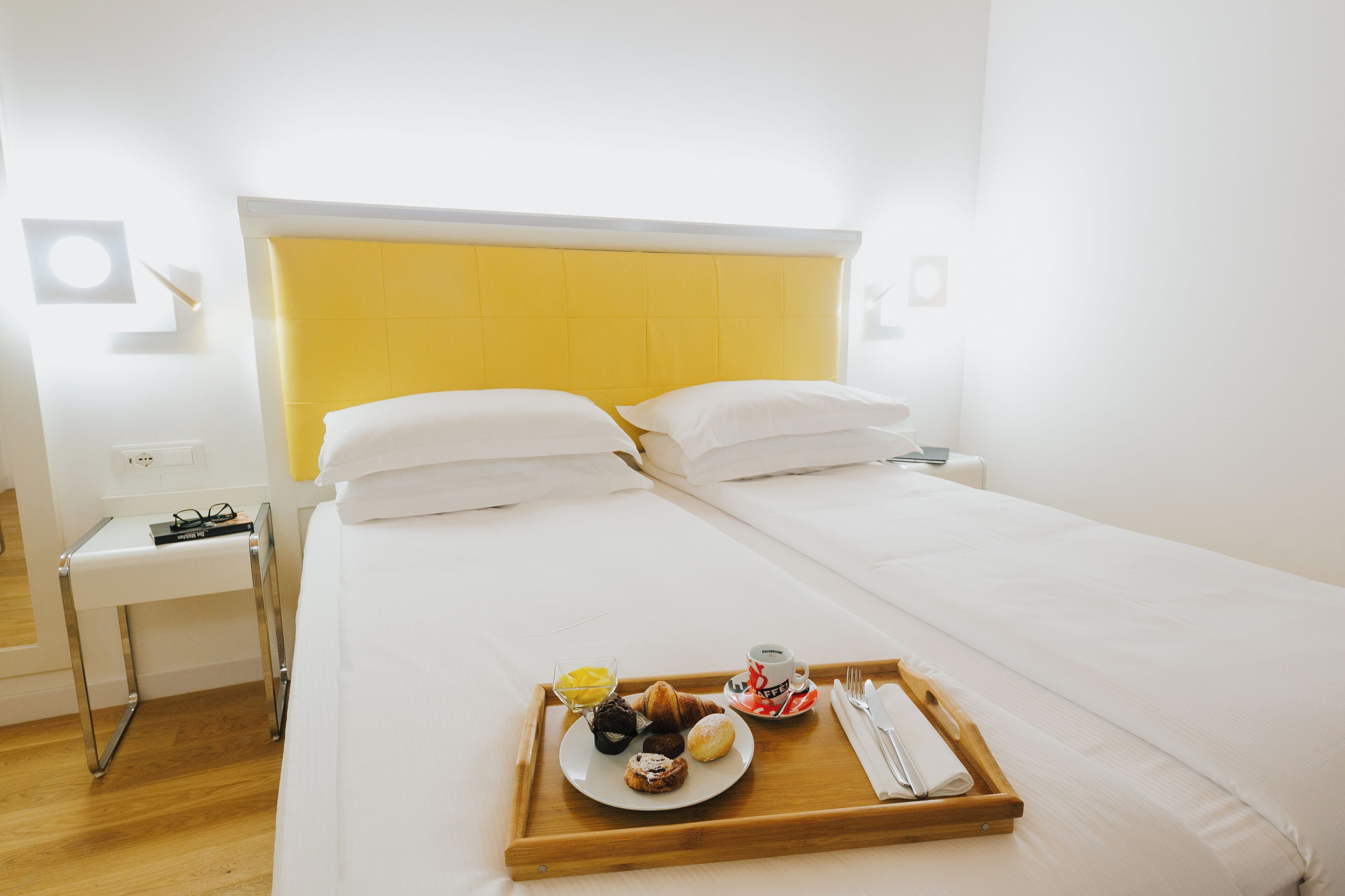 Quarto de hotel com uma cama, um tabuleiro com itens de pequeno-almoço e uma mesa de cabeceira com uma lâmpada.