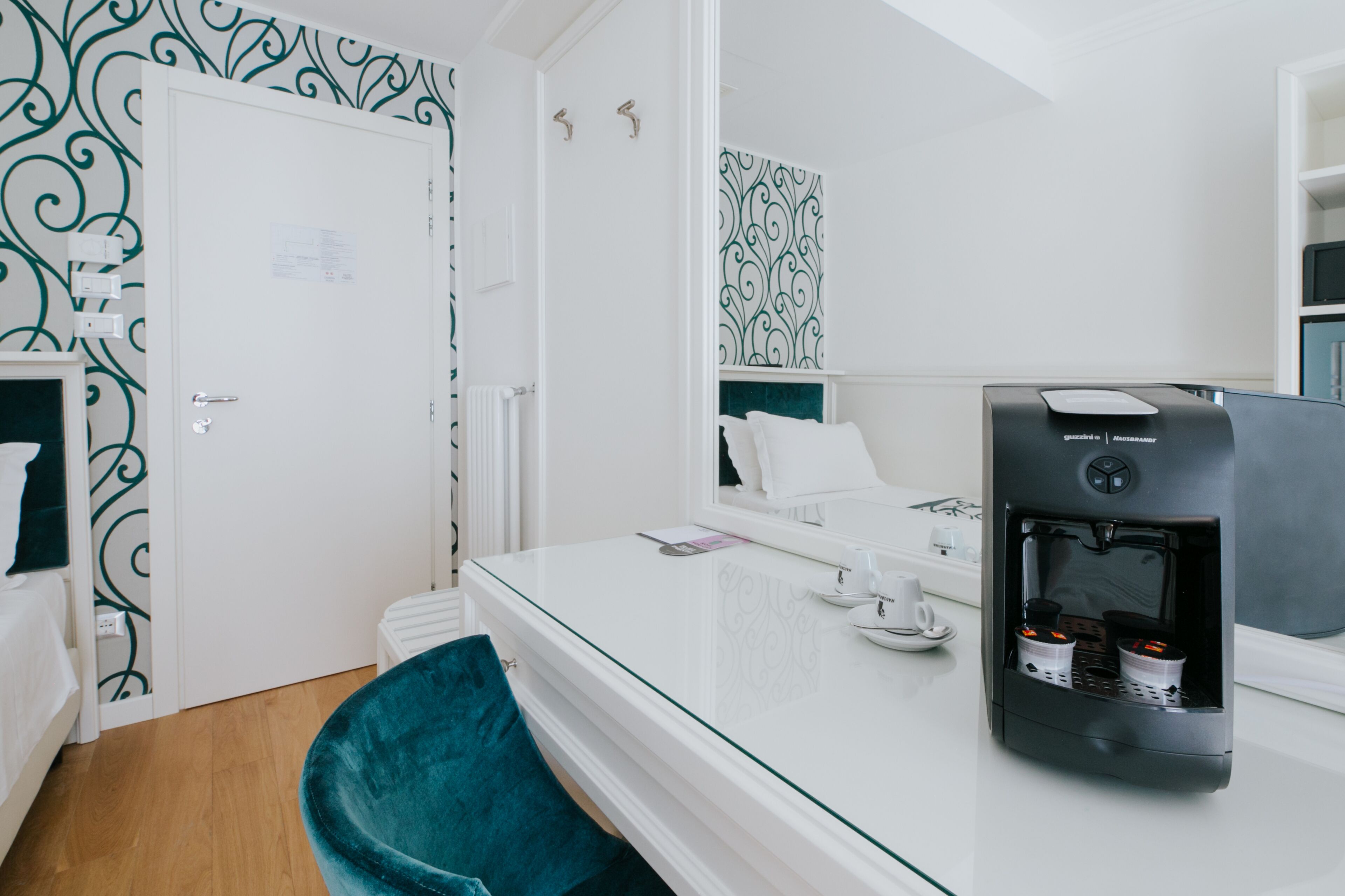 Une chambre d’hôtel moderne avec une table à plateau en verre, une machine à café noire, un fauteuil turquoise et un lit blanc.