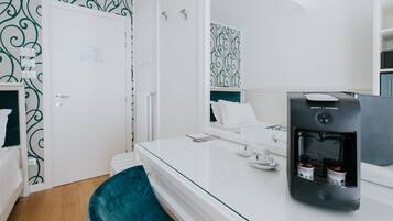 Une chambre d’hôtel moderne avec une table à plateau en verre, une machine à café noire, un fauteuil turquoise et un lit blanc.