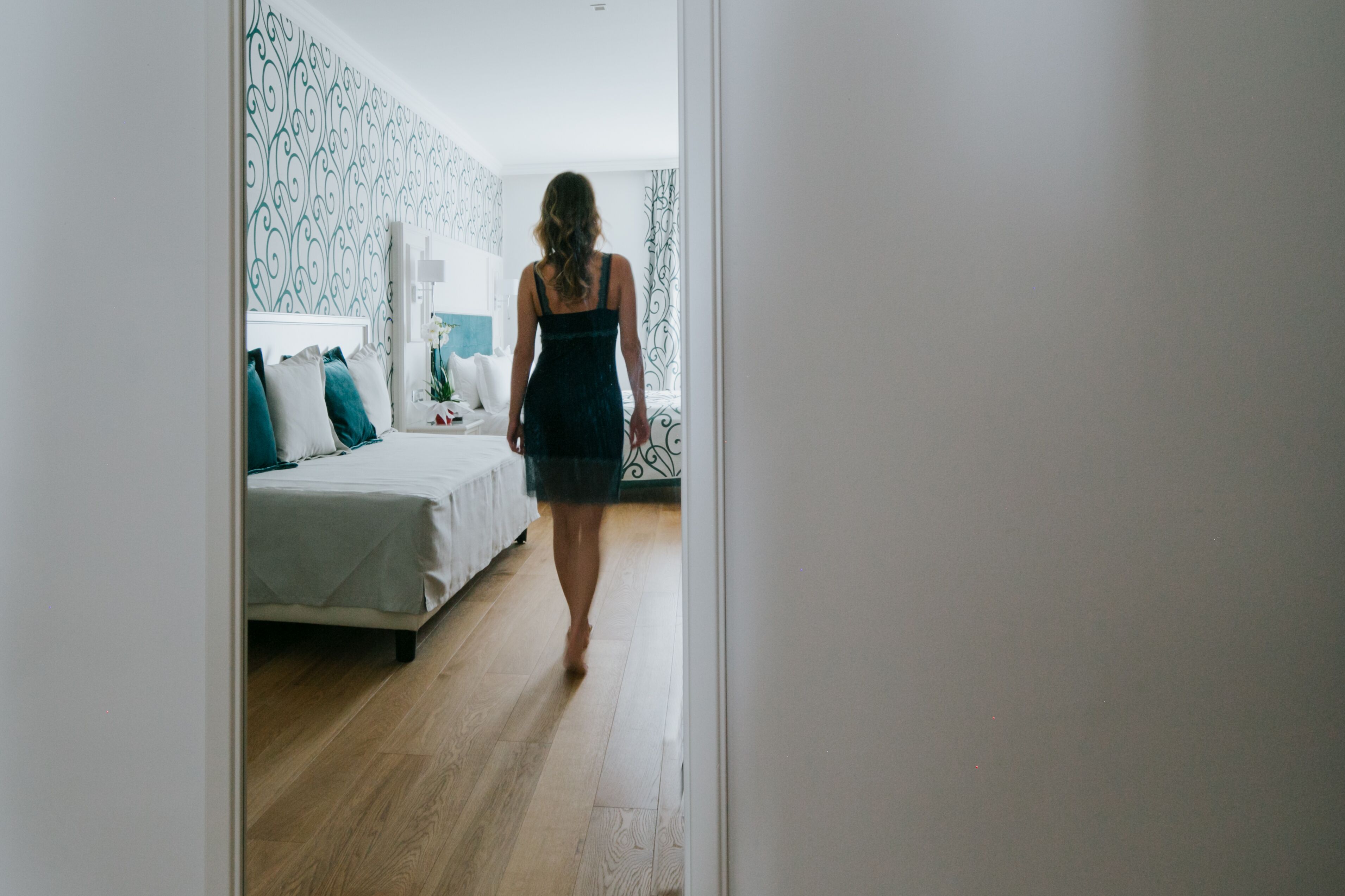 A woman walking through a hallway with a bed and a nightstand in the background.