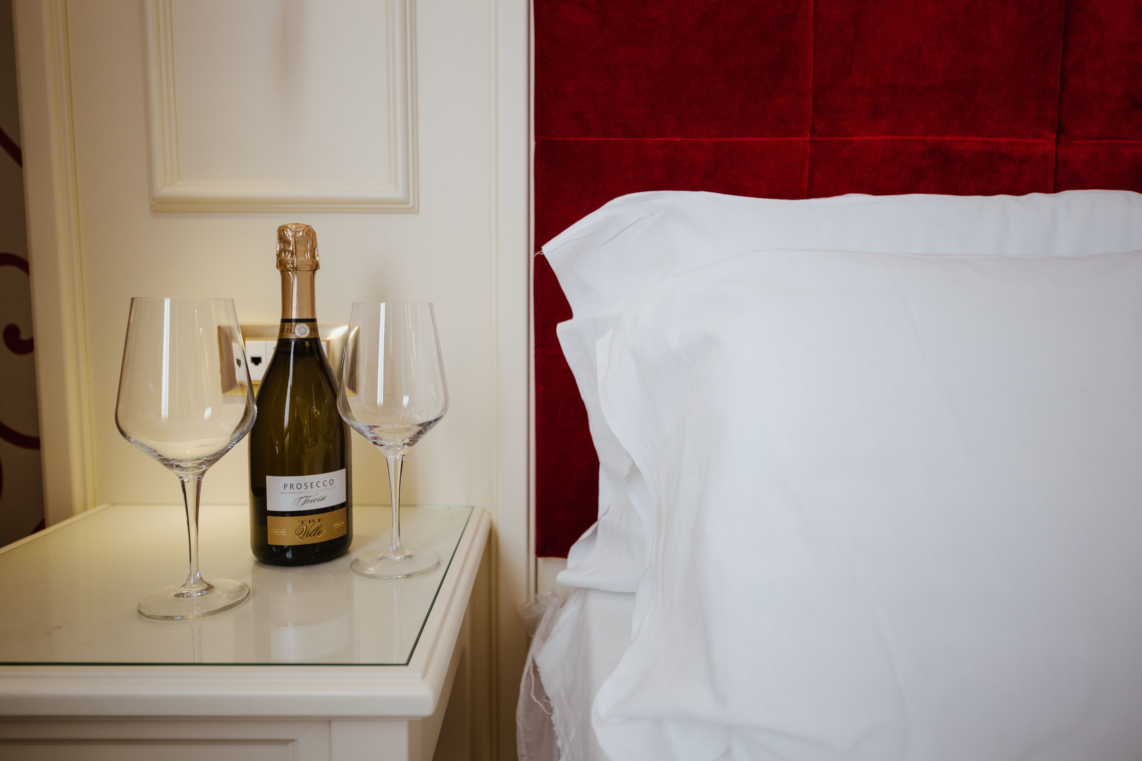 Quarto de hotel com cama branca, cabeceira vermelha e mesa com uma garrafa de Prosecco e duas taças de vinho.