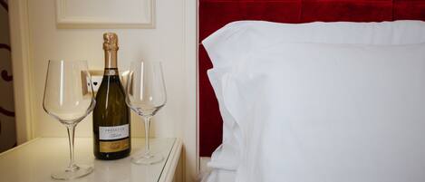 Quarto de hotel com cama branca, cabeceira vermelha e mesa com uma garrafa de Prosecco e duas taças de vinho.