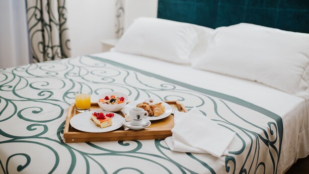 A neatly made bed with a green and white patterned bedspread, a tray with breakfast items including croissants, a bowl of cereal, and a glass of orange juice. - Hotel Città di Parenzo (Trieste)