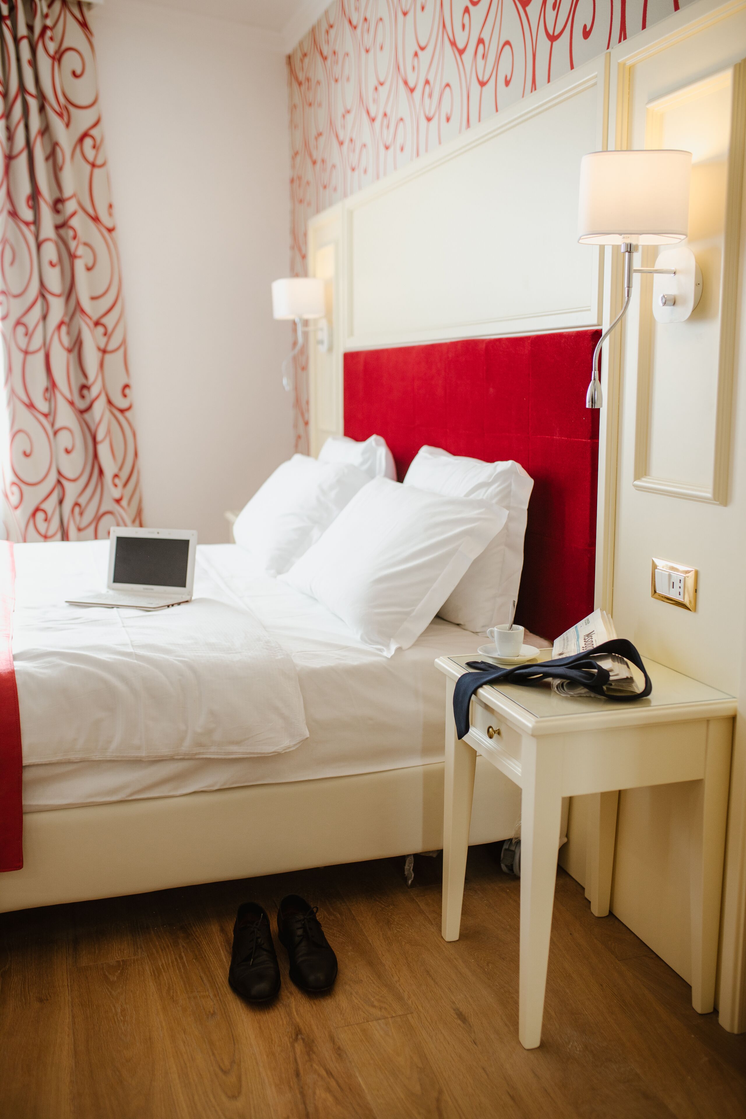 Ein Hotelzimmer mit einem Bett, einem Nachttisch, einer Lampe, einem Laptop und einem Paar Schuhen auf dem Boden.
