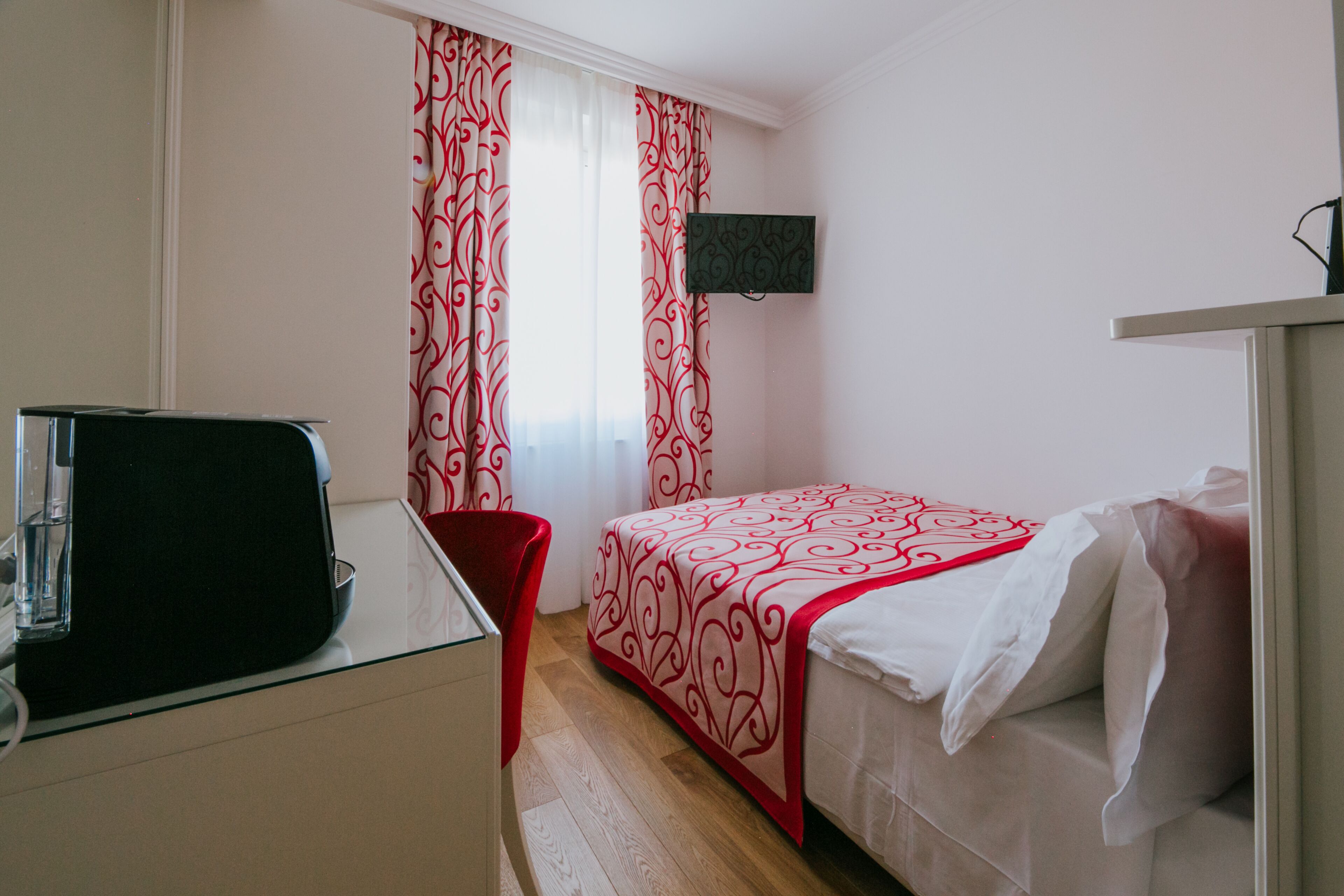 A hotel room with a bed, a desk with a chair, a TV mounted on the wall, and curtains with a red and white pattern.