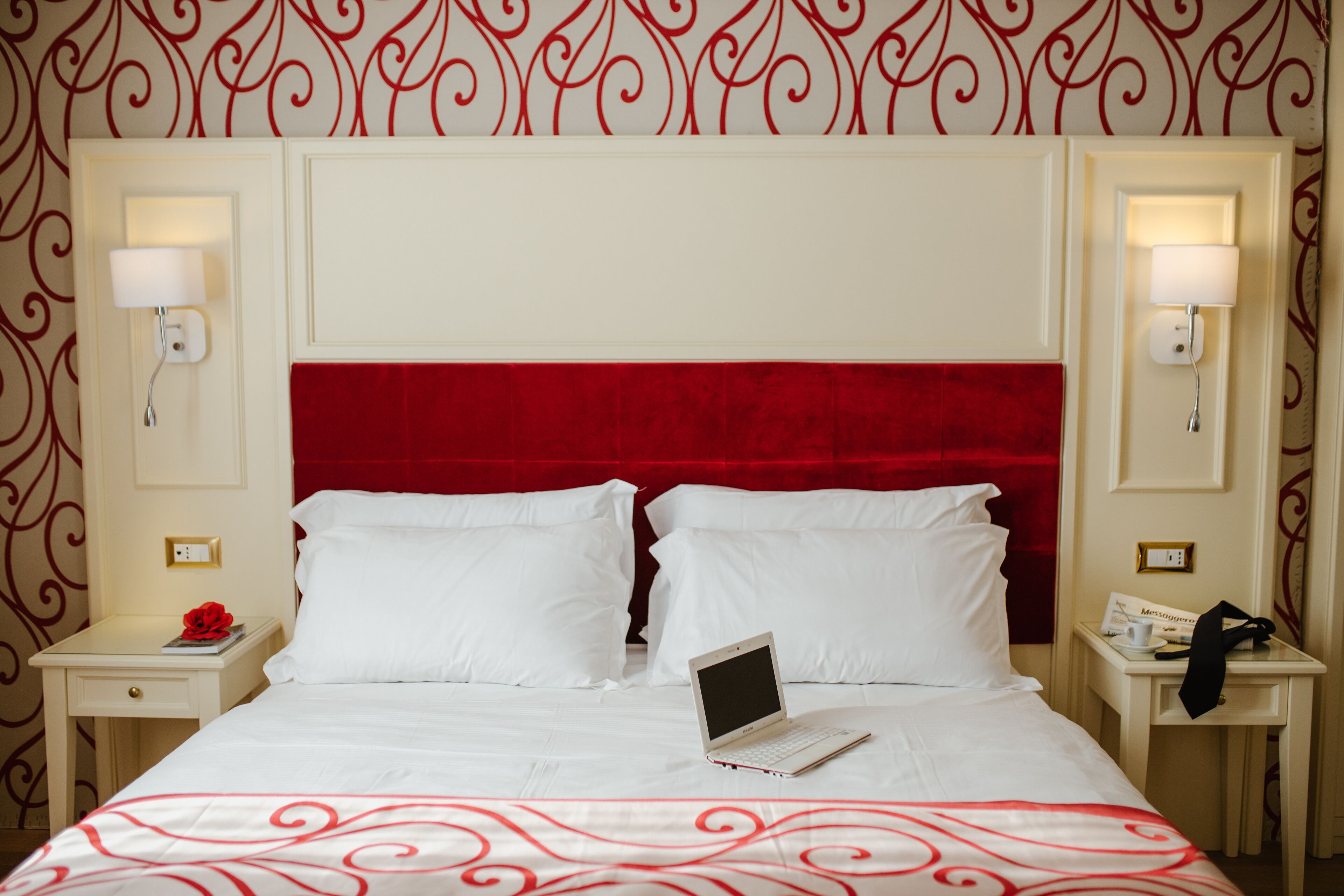 A hotel room with a red headboard, white bedding, a laptop on the bed, and two bedside tables with lamps and a phone.