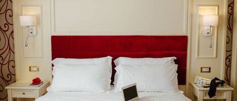 A hotel room with a red headboard, white bedding, a laptop on the bed, and two bedside tables with lamps and a phone.