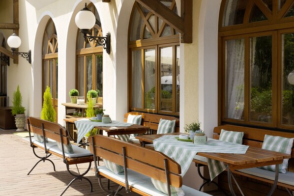 An outdoor seating area with wooden tables and benches, surrounded by large windows and potted plants.