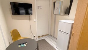 Una habitación pequeña con una televisión montada en la pared, un refrigerador, una silla y una mesa con un control remoto.