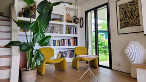 Uma sala de estar moderna com uma estante, duas poltronas amarelas, uma mesa de centro, uma grande planta verde e uma obra de arte emoldurada na parede.