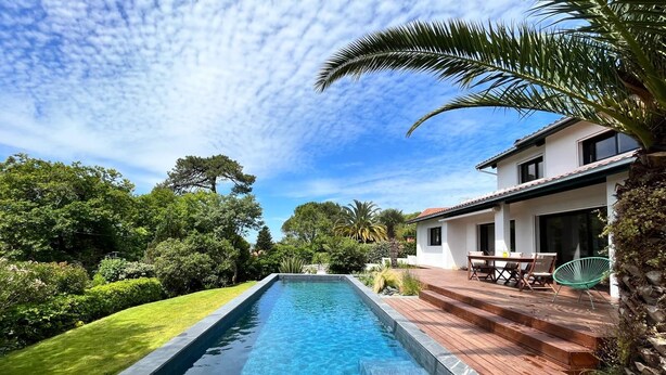 Piscina retangular com deck de madeira e borda de pedra, rodeada por vegetação exuberante e um edifício branco com telhado ladrilhado.