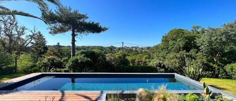 A rectangular swimming pool with a wooden deck in the foreground, surrounded by lush greenery and trees.