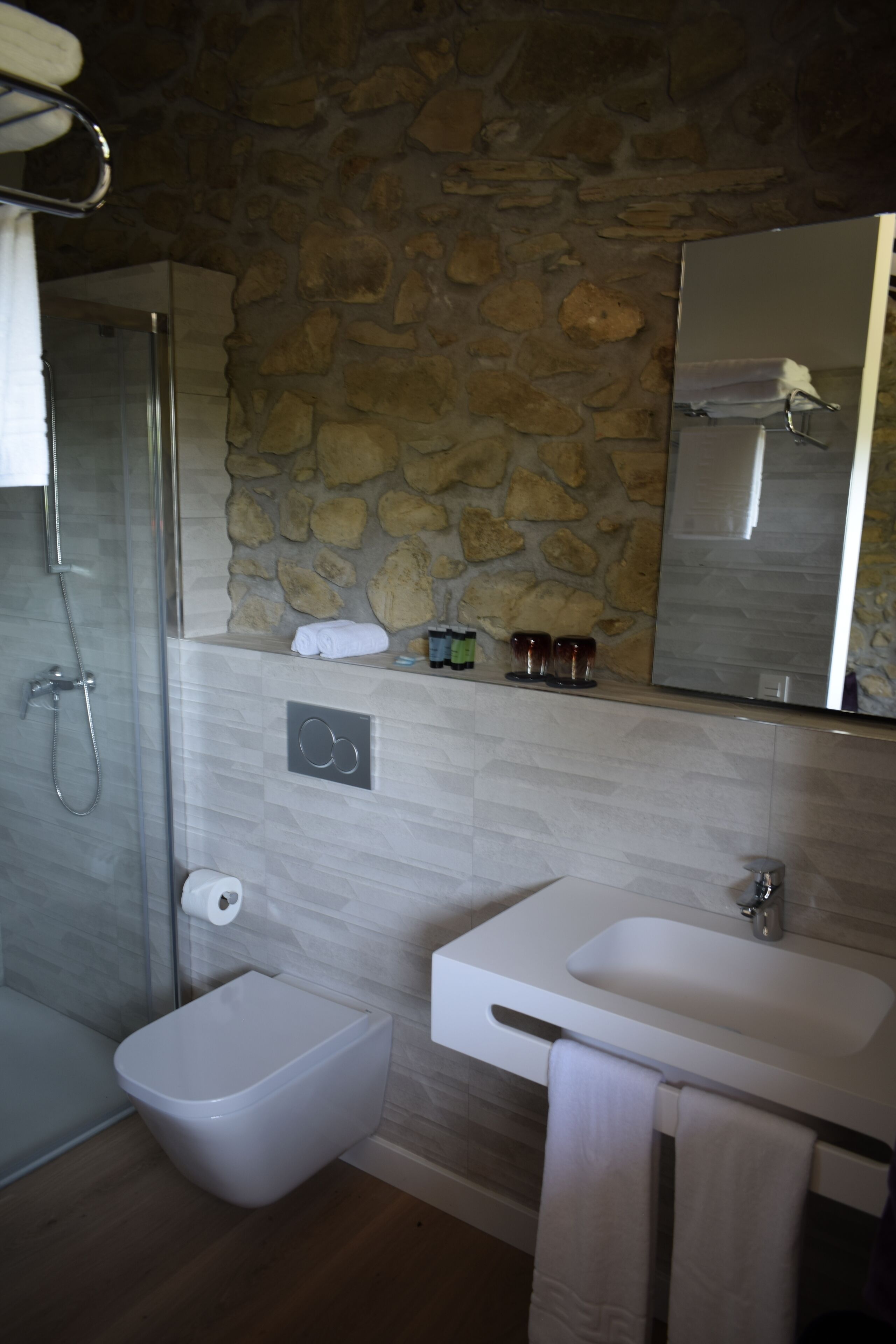 A bathroom with a stone wall, a glass shower enclosure, a toilet, and a sink with a mirror above it.
