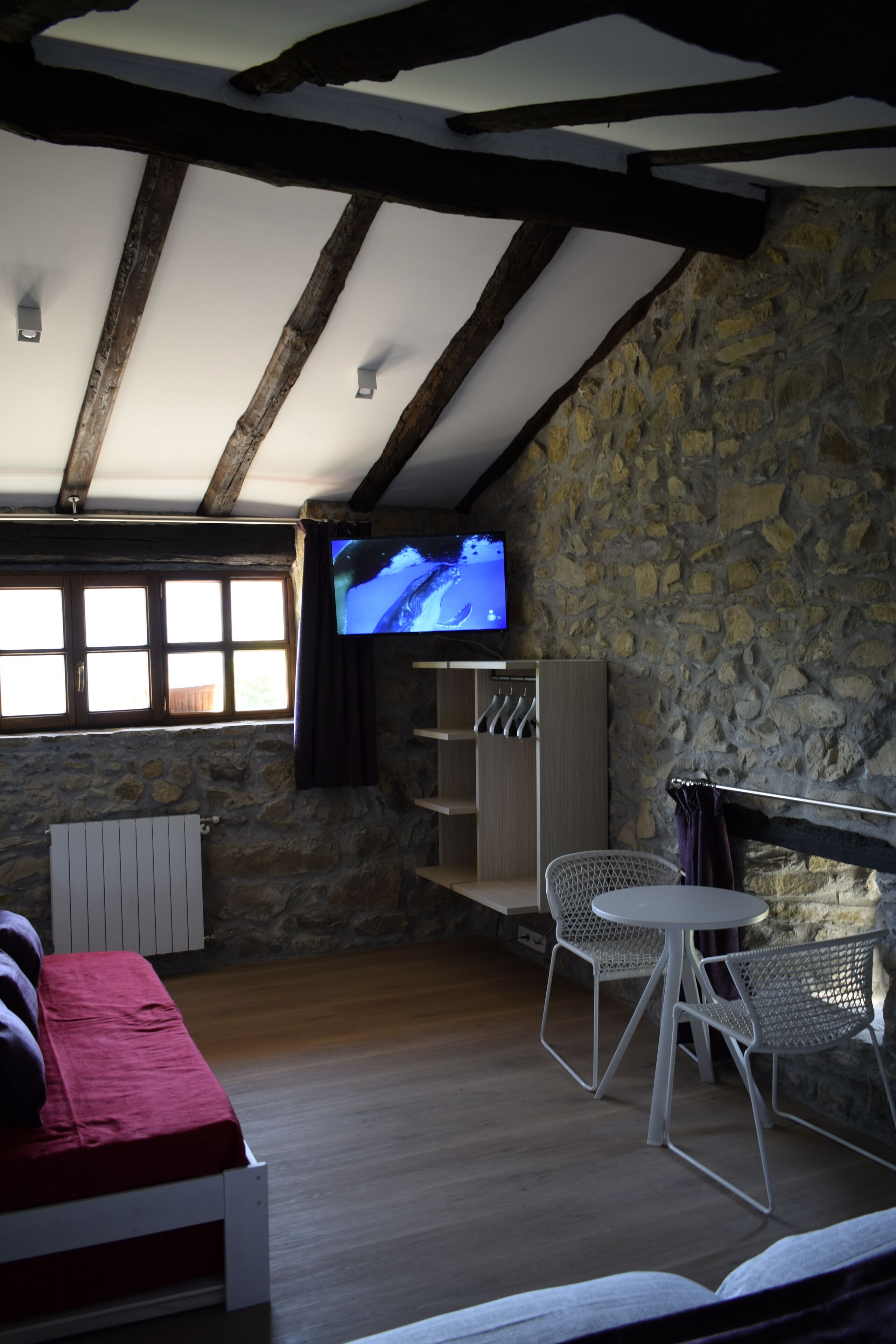 Ein gemütliches Zimmer mit Steinwand, Holzbalken, Fernseher, einem Bett und einem Tisch mit Stühlen.