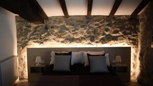 A bedroom with a stone wall, wooden beams, a bed with pillows, and bedside lamps. - Casa Rural Martiamuno Landetxea (Zumarraga)