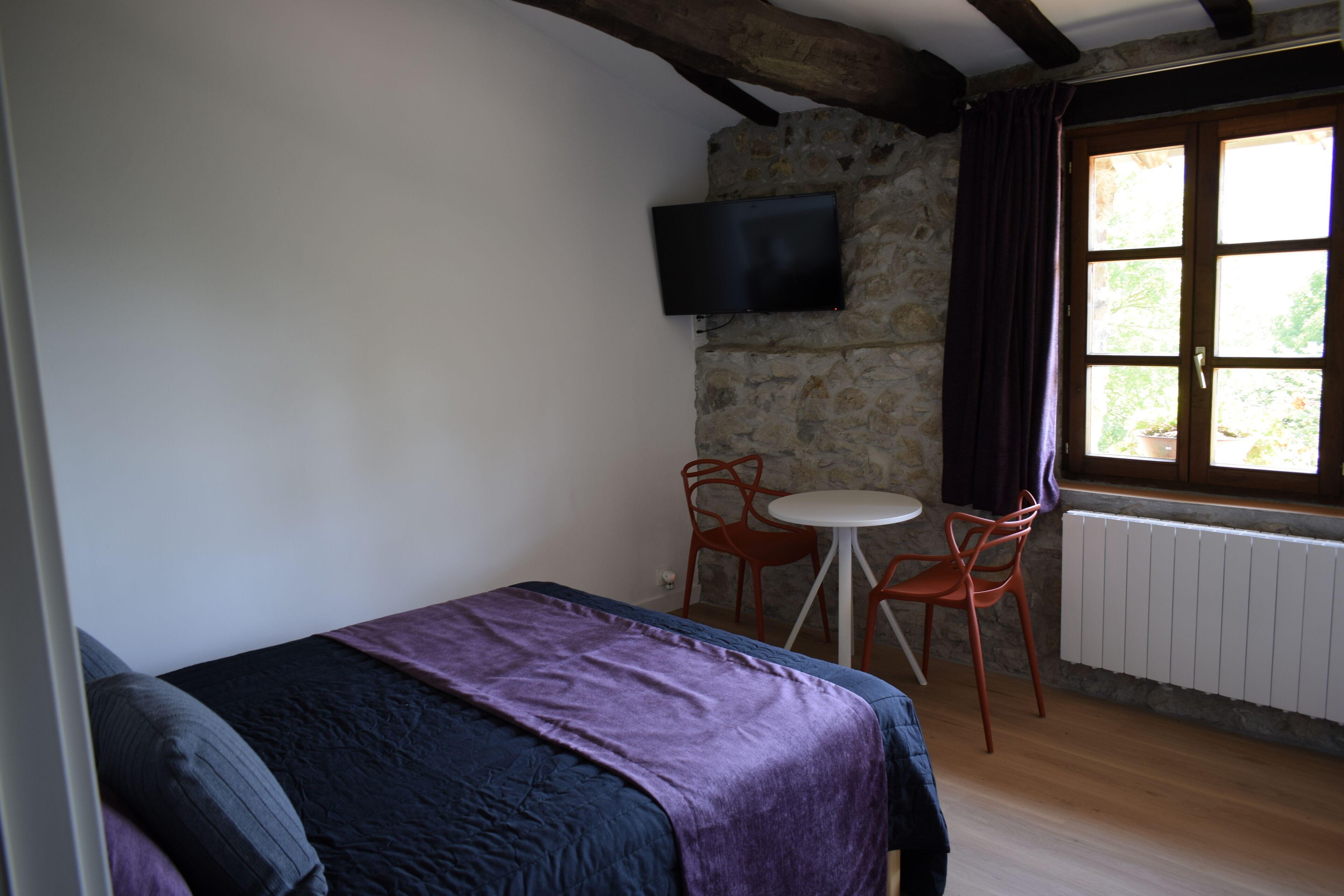 Un dormitorio con cama, televisor montado en una pared de piedra, una mesita con dos sillas y una ventana con cortinas.
