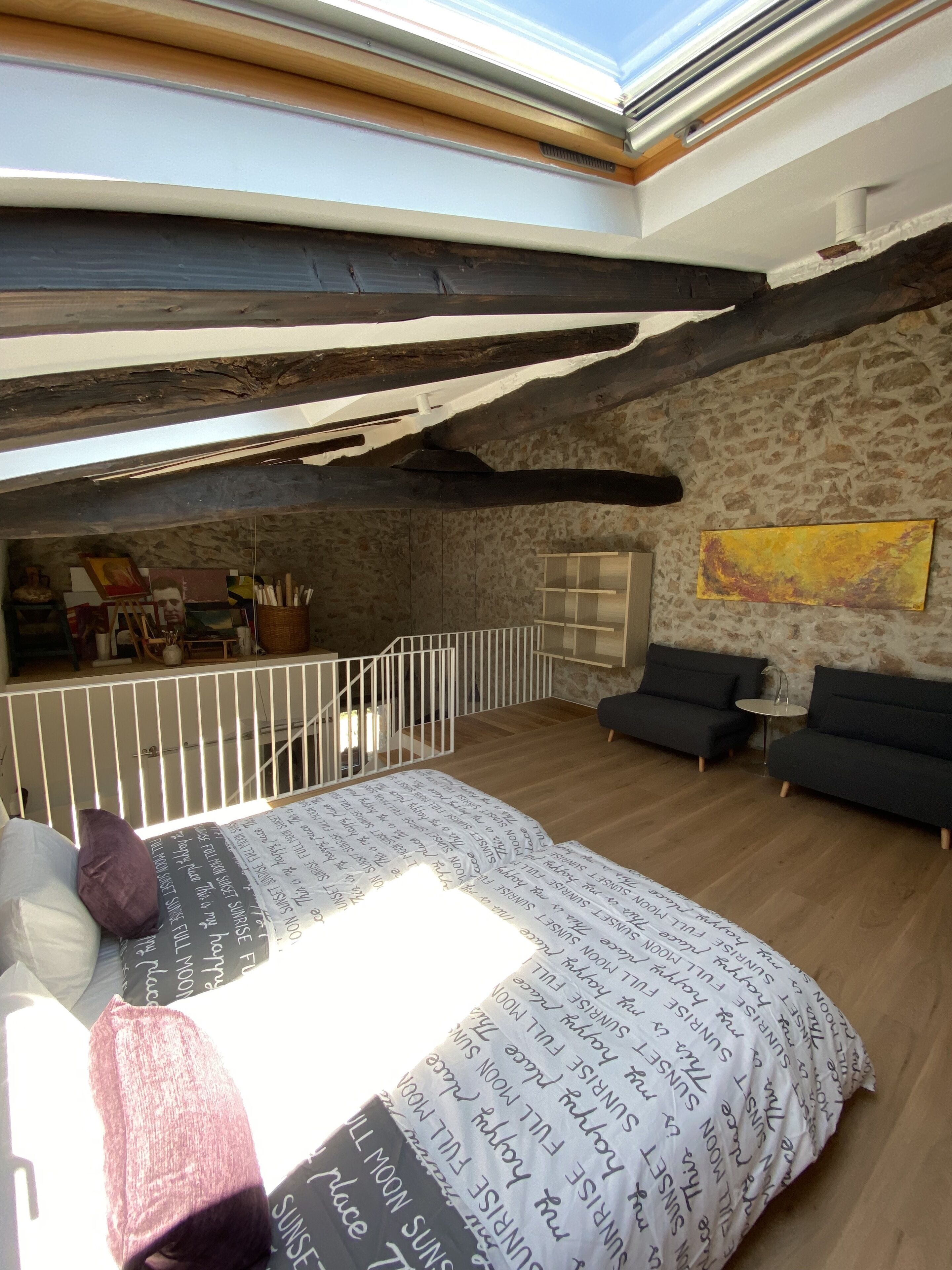 Un espacioso dormitorio tipo loft con techo alto, vigas de madera a la vista, pared de piedra y una cama grande con ropa de cama estampada.
