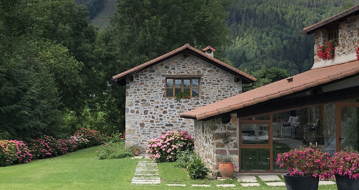 Een stenen huis met een pannendak, een glazen deur en een tuin vol roze bloemen.