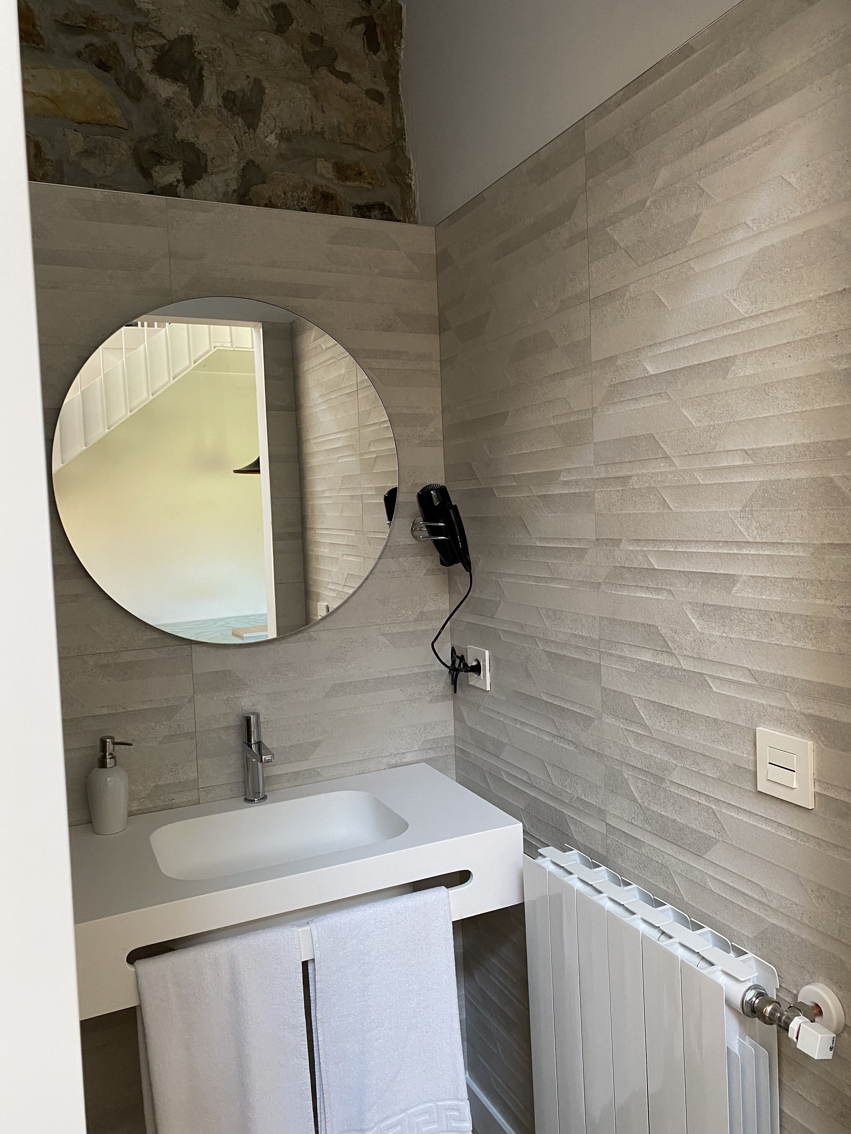 A modern bathroom with a stone wall, a round mirror, a white sink, a towel rack, and a radiator.