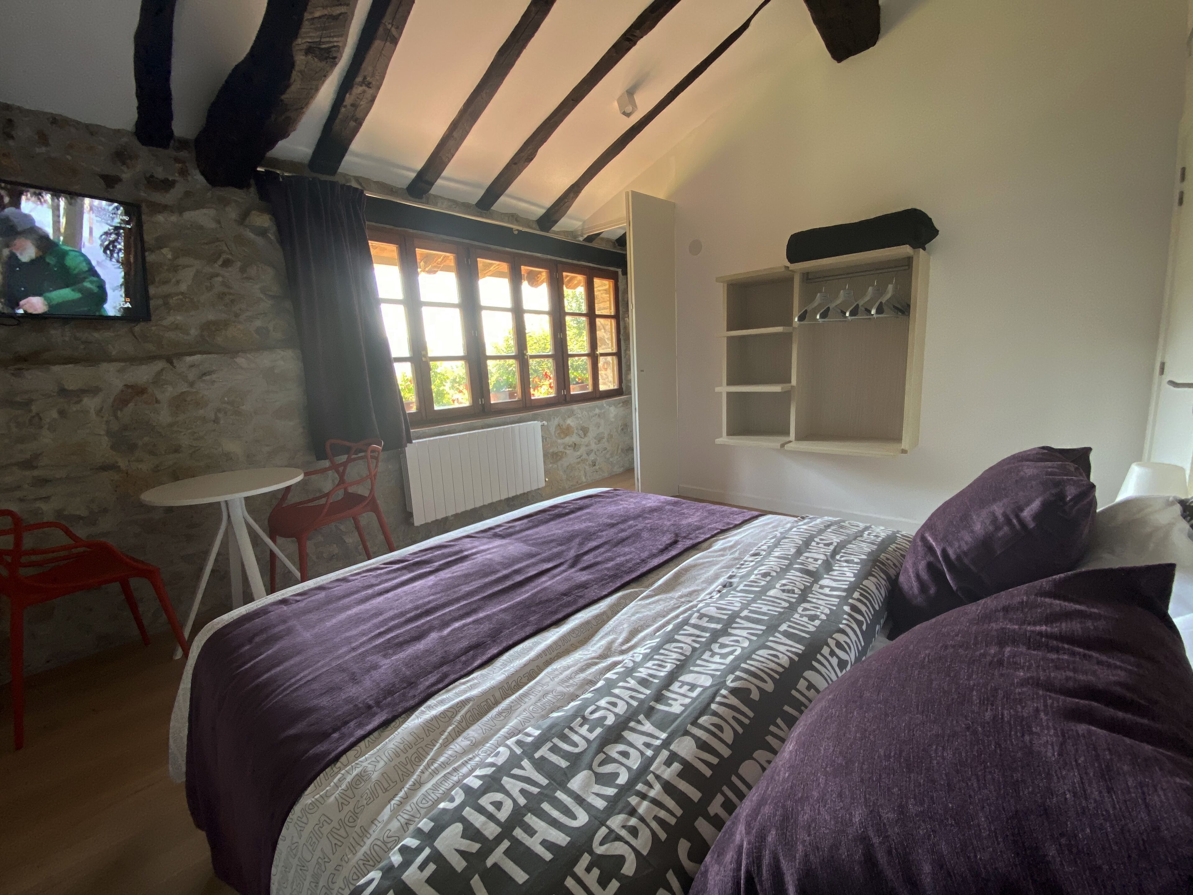 A bedroom with a bed, a small table, a chair, a window with curtains, and a stone wall.